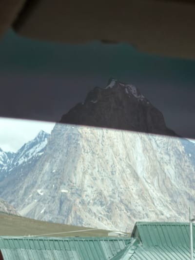 A mountainous landscape is depicted, with snow-capped peaks in the distance. In the foreground, there is a green, corrugated metal roof of a building. The image appears to be taken from inside a vehicle, with a dark interior element visible at the top. The location is Kurgiakh, India.