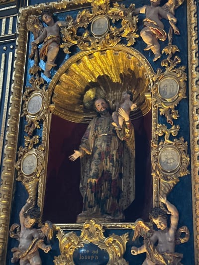 This media file showcases a highly ornate religious artwork, likely a section of an altarpiece or a large niche decoration, from Tepotzotlán, Mexico. The central subject is a statue of Saint Joseph, depicted with a beard and wearing a richly patterned robe adorned with floral motifs in red, green, and gold. He cradles the Christ Child in his arms. Behind Saint Joseph's head is a prominent, golden, rayed halo or mandorla, and the figures are set within a deep red recess.
The central scene is surrounded by an elaborate, multi-layered frame. The immediate border is gold, featuring intricate rococo-style scrollwork and foliage. Beyond this, on a dark blue background, are numerous sculpted putti (cherubic figures) in various dynamic poses, some with wings, integrated into the golden decorative elements. Several oval and circular medallions are also embedded within the golden scrollwork, each containing faint bas-relief imagery, possibly depicting saints or biblical scenes.
On the right side of the frame, within a golden cartouche, the Latin text "te ad Joseph" is visible, likely part of the phrase "Ite ad Joseph" (Go to Joseph). The artwork is characterized by its opulent use of gold, detailed carving, and vibrant colors, typical of Baroque or Churrigueresque style. The lighting suggests an indoor setting, and no specific activity beyond the static portrayal of the religious figures is occurring. The photograph is taken from a low angle, looking upwards at the artwork.