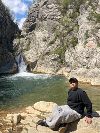 A partially rotated outdoor shot shows a young male sitting on a rock outcrop above a waterfall with a gorge to the left. The subject is wearing a black long-sleeved top, gray sweatpants, black sneakers, and a black baseball cap. His face is turned slightly towards the camera. He is partially illuminated. The waterfall cascades into a lower pool. The left side of the image features a tall, rocky cliff face with scattered vegetation and graffiti. The sky with a few clouds is seen in the top left corner. The setting is likely a natural area near Medven, Bulgaria.