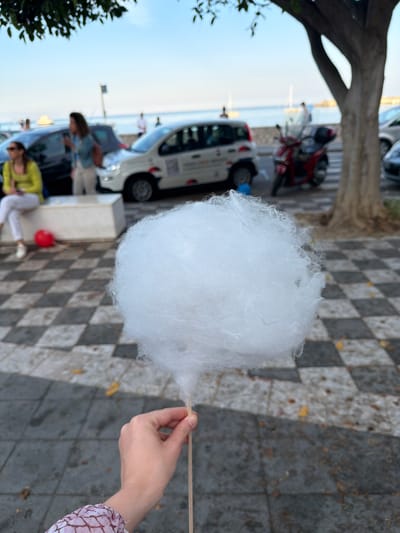 A close-up shot depicts a hand holding a large white ball of cotton candy on a stick. The person's arm is visible, clad in a pink and white patterned sleeve. The foreground features a black and white checkered sidewalk. In the blurred background, there are parked cars, people, and a tree. Based on the location and architectural details visible, the photo was taken on the lungomare of Chianchitta, Italy.