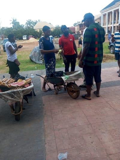 The image depicts an outdoor street vending scene in Ajalli, Nigeria.
In the foreground, a reddish-brown patterned paved surface with a scattered clear plastic bag is visible. To the left, a rusty wheelbarrow functions as a fruit display, containing items such as cut watermelon slices, bananas, and other produce, partially covered by clear plastic sheeting. A woman in a white t-shirt and dark skirt stands adjacent to this cart, holding and consuming a small, light-colored fruit.
Centrally, a second rusty wheelbarrow serves as a vendor's counter, holding black plastic bags and white sheeting. A barefoot woman in a blue and white patterned dress stands behind it, peeling a light-green fruit. She faces a man in a red t-shirt and dark cap, who holds a similar peeled fruit and looks towards her, indicating a transaction or interaction. To their right, a man in a green and black striped polo shirt and dark shorts stands with his back partially to the camera, wearing flip-flops and holding a dark object in his left hand. A fifth man, partially visible on the far right, wears a striped t-shirt and is eating a light-colored fruit.
The mid-ground features a strip of green grass separating the paved area from a dirt patch, where stacked concrete blocks are present. The background shows dense green trees and multiple buildings, including a prominent light-colored structure with several columns and a dark roof. A dirt road with various vehicles is visible further in the distance.