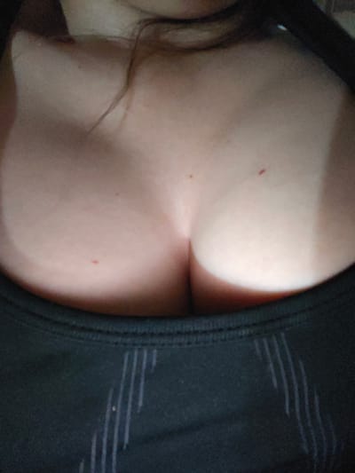The image shows a close-up view of a person's chest and upper torso, from the cleavage area up to the neck. The individual is wearing a black top with a patterned design. There are a few small marks on the skin, possibly moles or blemishes. The lighting creates a dramatic effect, highlighting the curves of the chest. The background is dark and out of focus, suggesting an indoor setting. There is no discernible information within the image that would allow for the identification of a specific location in Masally, Azerbaijan.