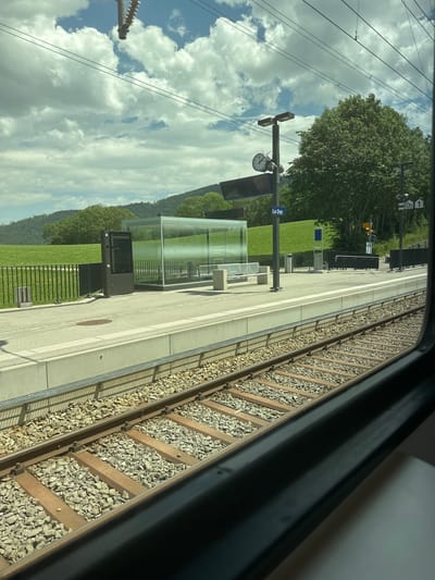 The image shows a train station platform at Le Day, Switzerland. The perspective is from inside a train, looking out. The platform has a modern glass shelter, benches, a trash can, and a ticket vending machine. A train track runs alongside the platform, with ballast and wooden sleepers visible. In the background, there is a lush green lawn, trees, and a hill covered in more trees. The sky is blue with scattered white clouds. A clock on a pole shows the time is a little after 9:00. Overhead power lines for the train are also visible. There are signs at the station, including one with "05 1" and another that reads "Le Day." The sun is shining, suggesting it's daytime. No people are visible in the image.