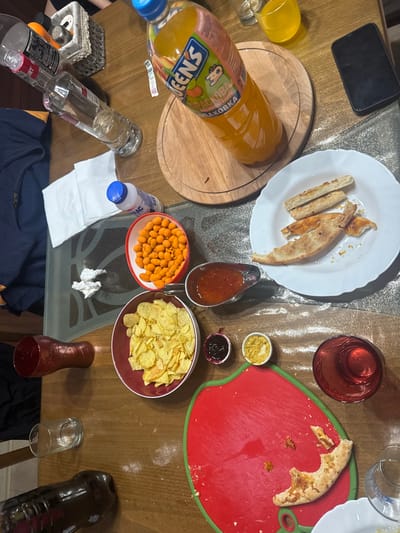 The image displays an overhead view of a wooden table covered with food and beverages, suggesting a casual gathering. No individuals are visible.
Objects on the table include:
* A large clear plastic bottle of "QUEENS" brand orange juice on a round wooden cutting board.
* A clear glass bottle of clear liquid, resembling spirits, positioned left of the juice bottle.
* A small white plastic bottle with a blue cap, labeled "КМС" or similar, adjacent to crumpled white napkins.
* A white rectangular plate containing several pieces of partially eaten pizza crusts or flatbread.
* A red bowl filled with orange-colored, coated peanuts.
* A red bowl filled with yellow ridged potato chips.
* A silver gravy boat holding a quantity of reddish-orange sauce.
* Two small white ramekins containing a dark red jam-like substance and a yellowish condiment, respectively.
* A red, apple-shaped cutting board with green trim, displaying crumbs and a piece of partially eaten pizza crust.
* Various drinking glasses: a clear glass with some liquid, a red opaque glass, a red clear glass with red liquid, a yellow clear glass with yellow liquid, and another clear glass.
* A black smartphone is placed face down near the juice bottle.
* A silver, textured shaker or container is visible in the upper left corner.
* A dark blue piece of fabric, possibly clothing, is partially visible on the left side.
The table surface appears to be wood, with a transparent patterned covering over a portion of it. The overall scene indicates recent food and drink consumption.
The image does not contain any visual identifiers or markers to confirm the location as Kanchevo, Bulgaria.