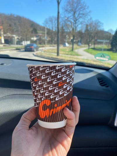 A right hand holds a brown, corrugated paper cup displaying the 'Grimmiger' logo in white and orange lettering, positioned within the interior of a vehicle. The cup's contents are not discernible. Beyond the vehicle's windshield, a clear day illuminates a road intersection in Bensheim, Germany. A dark sedan is observed moving away from the viewer on the road. The environment features bare deciduous trees, green ground cover, and road signage, including a blue circular sign and a green oval sign. Distant hills, covered in leafless trees, form the horizon under a bright blue sky.