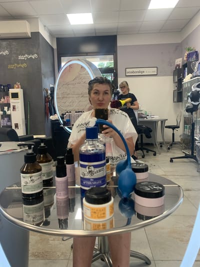 A woman with dark hair, wearing a white smock and blue gloves, is taking a selfie in a mirror. She is in a brightly lit salon, likely a barbershop or hair salon, with tiled floors and grey walls. Several bottles of grooming products, including "Old Barber" brand shaving gel and aftershave, are arranged on a round silver table in the foreground. In the background, another woman with blonde hair and glasses is visible, holding a yellow hair dryer. The salon appears to be located in the city of San Quirico d'Orcia, Italy, as evidenced by signage on the wall that reads "San Quirico d'Orcia."