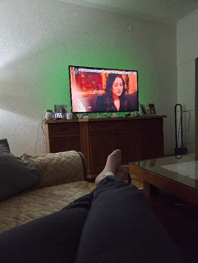 The image presents an interior residential scene in Negorci, North Macedonia. The perspective is low, originating from a reclining position. In the immediate foreground, a person's lower legs, clad in dark trousers, are extended forward with feet crossed at the ankles. Light-colored socks, one with a red detail, cover the feet. To the left, a section of a light brown, quilted sofa with a dark gray throw pillow is visible. To the right, a wooden coffee table with a glass surface is present.

Centrally positioned is a flat-screen television displaying a medium close-up of a seated female with dark hair. The screen includes overlaid text at the top and bottom. Beneath the television, a dark wooden cabinet with multiple drawers supports several objects, including a digital clock indicating "17:55," various small decorative items, and framed photographs. Green backlighting behind the television illuminates the textured wall surface directly behind it. The surrounding walls are textured and predominantly light-colored. A white ceiling with crown molding is visible at the top. On the far right, a dark, vertical floor lamp or similar fixture is partially obscured. On the far left, a portion of another wooden cabinet is visible behind the sofa.