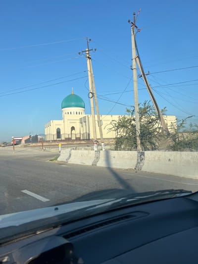 The image shows a view from inside a vehicle looking out towards a mosque with a teal-colored dome, located in Yangi Chinaz, Uzbekistan. The mosque is a light-colored, modern building with arched windows and a fence in front of it. It is surrounded by some vegetation and utility poles with many wires. The sky is clear and blue, and the road in the foreground has a white line marking. The perspective suggests the vehicle is moving on a road adjacent to the mosque.