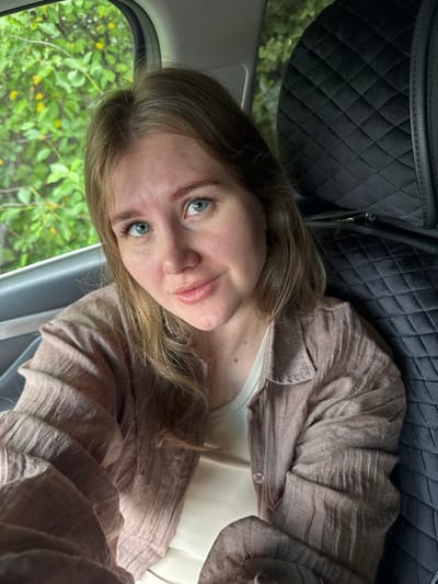 A woman with blonde hair and blue eyes is seated in the back of a car. She is wearing a light brown, crinkled shirt over a white tank top. To her left, through the car window, a lush green foliage is visible with hints of yellow flowers. To her right is the quilted, black headrest of the car seat. The image is taken from a close-up, frontal perspective, with the woman looking directly at the camera. The location is specified as Bolshiye Kusty, Russia.