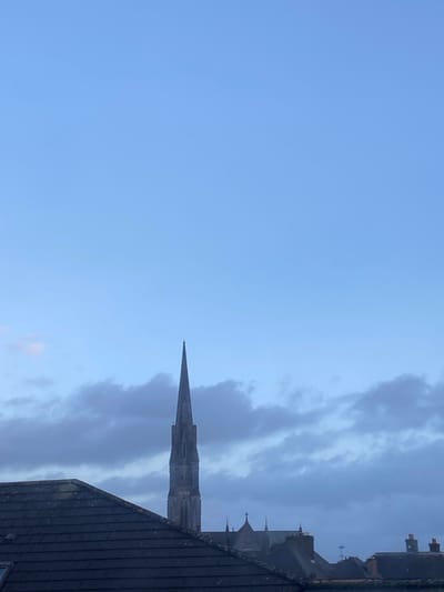 The image displays a distant view of St. Mary's Cathedral in Limerick, Ireland, specifically its prominent stone spire. The spire, with its multi-tiered architectural detailing and sharp pinnacle, is centrally positioned, rising above the dark-tiled roof of a foreground building, which occupies the lower quarter of the frame. Beyond this immediate rooftop, additional building structures with pitched roofs and visible chimneys are present in the mid-ground, partially obscuring the lower sections of the cathedral. The upper two-thirds of the image consist of a light blue sky with scattered, soft-edged grey stratocumulus clouds, suggesting an overcast or twilight condition. No individuals, specific actions, or interactions are discernible within the scene.