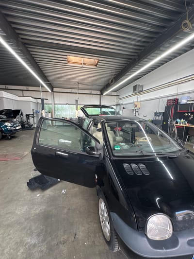 In an auto repair shop in Oberschleißheim, Germany, a black Renault Twingo is parked. The car's driver-side door is open, and the rear hatch is also ajar. The interior of the car, including the beige leather seats, is visible. In the background, another car, a dark green Fiat 500, is on a lift with its hood open. Tools and equipment are scattered throughout the workshop, and fluorescent lights illuminate the space from the corrugated metal ceiling. The floor is concrete and shows signs of wear.