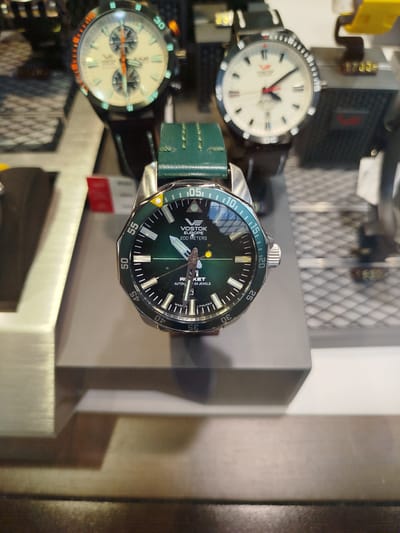 A Vostok Europe watch with a green band is displayed in a store in Marki, Poland. The watch has a green dial with white luminous markers and hands, and a rotating bezel with minute markings. The watch is placed on a gray stand. In the background, other watches and display cases are visible, as well as some price tags. The lighting is bright, suggesting an indoor retail environment.