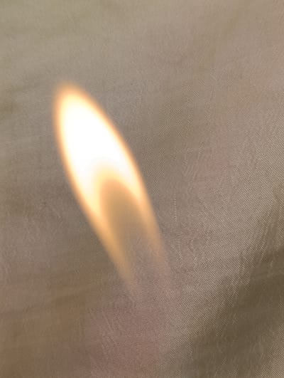 The media file depicts a close-up, abstract view of a textured, light brown fabric illuminated by a bright, flickering light source. The light is predominantly white and yellow, with softer orange and pink hues, suggesting a flame or a strong, warm light reflecting off the surface. The fabric itself has a visible woven texture, appearing to be a tightly woven material like canvas or a durable synthetic fabric. The light casts a soft, elongated glow across the fabric, creating a dynamic interplay of light and shadow. There are no discernible people, specific objects, or a clear scene or setting. The activity taking place is implied by the light source, which could be a candle, a gas burner, or a similar flame. There are no location cues beyond the fabric's texture. The overall impression is one of warmth and light against a neutral, textured background. No text is visible in the image.