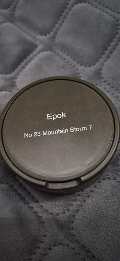 The image is a close-up shot of a circular, dark-colored object, likely a lid or tin, resting on a textured gray surface. The object has text inscribed on it. The text reads "Epok" in a larger font, and below it, "No 23 Mountain Storm 7" in a smaller font. The object is positioned at an angle, with its textured lid facing the viewer. The background surface appears to be a soft, quilted fabric, possibly a blanket or a piece of furniture. The lighting suggests an indoor setting, and no specific time of day or weather conditions can be inferred. The overall impression is that of a product shot, highlighting the branding and product name. There are no people visible in the image, and the focus is entirely on the object and its inscription. The color palette is muted, consisting of grays and dark tones, contributing to a simple and uncluttered aesthetic. The location context of Lillehammer, Norway, does not provide any immediate visual cues within the image itself, but it might be relevant to the product being displayed.