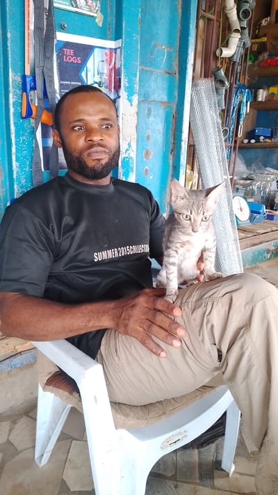 The image shows a man seated in a white plastic chair with a grey tabby kitten perched on his lap. The man is wearing a black t-shirt with the words "SUMMER 2015 COLLECTION" printed on it and beige pants. He has dark hair and a beard. The kitten is looking forward with its ears perked up.
Behind the man is a blue, textured wall with a sign that reads "TEE LOGS LEANING SERVICES." To the right of the man is a cluttered area with various objects including pipes, tools, and what appears to be a metal mesh roll. There is also a weighing scale visible.
The scene appears to be a shop or workshop environment. Given the information provided, the location is identified as Asubi, Nigeria.