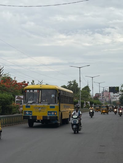 A bright yellow school bus, identified by the "R.G.M." lettering on its windshield, is the main subject of this image, driving on a paved road. The bus is an Ashok Leyland model and displays a license plate reading "AP21T X7682". The bus is in motion, as indicated by the blurred background.
Other vehicles are also present on the road, including auto-rickshaws and motorcycles. A man on a dark-colored scooter is in the foreground, also in motion. Behind him, several auto-rickshaws and motorcycles are visible, suggesting moderate traffic.
The environment appears to be an urban or suburban setting in Kurnool, India, characterized by streetlights, buildings in the distance, and trees lining the roadside. The sky is overcast with clouds. The road surface shows some imperfections, like small cracks and patches. The scene captures a typical day on a busy road in the city.