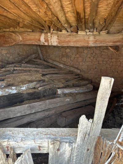 The image shows the interior of a rustic structure, likely an old barn or shed, located in Alpuente, Spain. The scene is dominated by raw, weathered wood and stone construction.

The ceiling is made of rough-hewn wooden beams and smaller branches, creating a rustic and uneven surface. A large, thick beam runs horizontally across the middle of the image, supporting the ceiling structure and resting against a stone wall. This beam shows signs of wear and discoloration.

The back wall is constructed of rough-cut stones, laid in an irregular pattern. Piles of chopped logs and branches are stacked against this wall and fill much of the floor space. Some of the wood appears to be partially covered by dry straw or hay. The logs vary in size and condition, with some appearing freshly cut and others weathered and aged.

In the foreground, more wooden beams and planks are visible, some of which appear to be part of a damaged or decaying structure, possibly a fence or partition. These foreground elements are also made of aged, greyed wood, some showing signs of splitting and damage. Small, rusty nails are visible in some of these wooden pieces.

The overall impression is one of disuse and decay, a scene of agricultural storage or a place of shelter that has fallen into disrepair. There are no people or animals present in the image. The lighting suggests it is daytime, with natural light filtering into the interior, creating shadows and highlighting the textures of the materials.