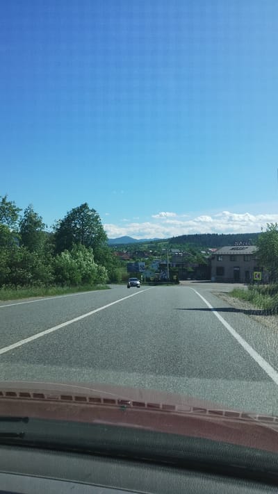 The image depicts a road in Deliatyn, Ukraine, from the perspective of a vehicle's interior. The road stretches ahead, with a white line marking the edge. Buildings are visible along the road, and a forested hillside rises in the background beneath a partly cloudy sky. Another car is visible driving on the road. A portion of the dashboard and windshield is visible in the foreground.