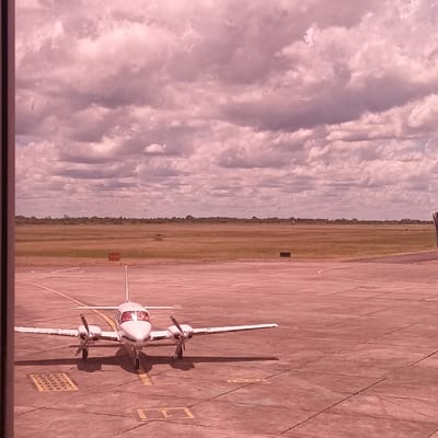 A white turboprop aircraft with red accents is parked on an airport tarmac. The plane faces towards the horizon. Beyond the tarmac is a grassy area bordering a field. The sky is filled with dense, pink-tinged clouds. The tarmac has painted markings and appears to be made of concrete. The lighting suggests it might be late afternoon. The perspective indicates the photo was taken from an elevated position, possibly inside an airport terminal.