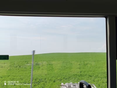 The image captures a wide, rolling green field under a bright, overcast sky. The foreground is framed by the interior of a vehicle, with a dark grey trim at the top and a vertical pillar on the right. A silver pole with a small blue and white sign is visible in the middle ground, to the left of a dark green rectangular sign. A stack of wooden pallets is present in the bottom right corner. The overall scene depicts a rural landscape.