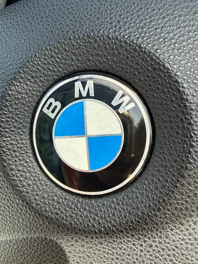 The image displays a close-up view of the BMW logo on what appears to be a steering wheel. The logo is circular with a black background and a silver border. Inside the circle, the BMW emblem is visible, consisting of four quadrants: two blue and two white. The letters "B," "M," and "W" are arranged in silver above the emblem. The surrounding surface is textured, likely leather, and exhibits a dark gray color, consistent with a car interior. The lighting suggests an indoor setting with natural light casting shadows. The location is described as Fritzlar, Germany, though the image itself does not provide explicit contextual clues to confirm this specific location. There are no people or actions depicted in the image.