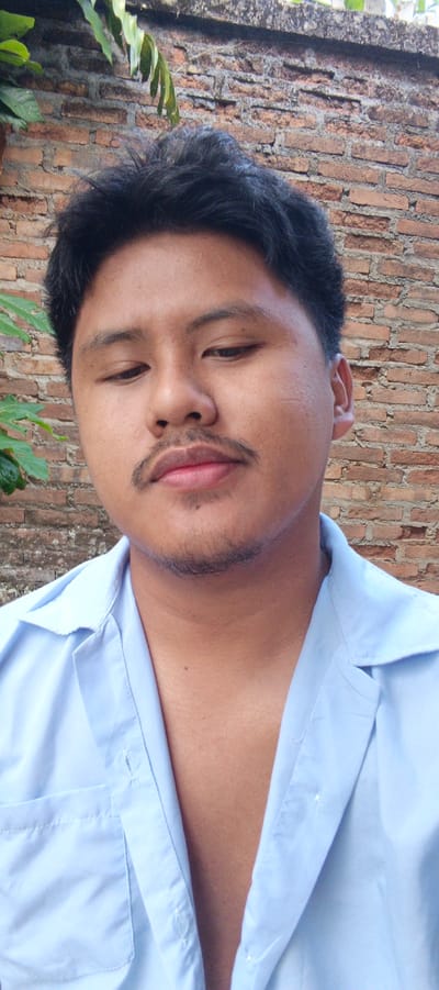 A medium shot image shows a young man with dark hair and a mustache, wearing a light blue collared shirt. He is positioned in front of a brick wall, with some green foliage visible in the upper left corner. The man is looking downwards and to his left. The scene appears to be outdoors. The location is in the city of Kemuning, Indonesia.