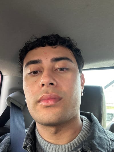 A close-up, chest-up portrait depicts an adult male inside a vehicle, located in Neuilly-Plaisance, France. The subject, centrally framed, has dark curly hair, brown eyes, and full lips. A small reddish blemish is visible on his forehead. He wears a small silver hoop earring in his left ear. His clothing consists of a grey ribbed knit sweater underneath a dark grey denim jacket. A blue seatbelt strap passes over his left shoulder, secured by a light grey buckle mechanism to the left of the frame. The vehicle's grey headliner is visible in the upper portion, and the top of a dark headrest is partially visible behind his right shoulder. Through the vehicle's window on the right, a blurry outdoor environment with indistinct structures is visible under ambient lighting conditions. The subject is looking directly into the camera.