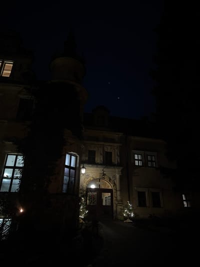A low-angle, dark image of a building at night. Illuminated windows punctuate the facade. One prominent window features a circular architectural detail above. Christmas lights are draped around it. A cylindrical tower-like structure appears to the left, extending towards the dark sky. The building is partially obscured by vegetation. Two distinct points of light, potentially stars, are visible in the upper left corner.