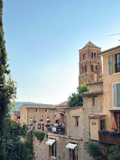 The image depicts a view of the historic village of Moustiers-Sainte-Marie, France, under a clear blue sky. A prominent stone bell tower, characteristic of Provençal churches, rises in the background, adorned with arched openings and a cross at its apex. In the foreground and mid-ground, traditional stone and stucco buildings with terracotta tile roofs line a street. Some buildings feature outdoor seating areas with tables and chairs, suggesting cafes or restaurants. A tall evergreen tree is visible on the left side of the frame, and lush greenery cascades down a stone wall. Small details like window shutters, outdoor lighting fixtures, and a pink umbrella on a balcony add to the charm of the scene. A few people are seated at an outdoor table on a balcony, engaged in conversation. The overall atmosphere is serene and picturesque, capturing the essence of a charming European village.