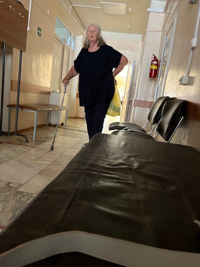 An elderly woman with gray hair stands in a hospital hallway, holding a cane in her right hand and a yellow plastic bag in her left. She is wearing a dark blue top and dark pants. The hallway is lined with pale yellow walls and has tiled flooring. To the left, there is a wooden coat rack with hooks and a bench. To the right, there are several dark brown chairs lined up along the wall, and a red fire extinguisher mounted on the wall. A doorway is visible behind the woman, leading to another room. The scene appears to be a waiting area in a medical facility in Ikryanoye, Russia.