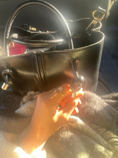 The scene depicts a close-up interior view, likely within a vehicle, located in Monastir, Tunisia. A fair-skinned human right hand, wearing a gold-colored ring with a white butterfly ornament on the ring finger and a simple gold-colored band on the middle finger, is centrally positioned, illuminated by strong, warm sunlight from the left. The hand is holding a black knotted drawstring with a silver-colored metallic end. This drawstring is part of a black, structured handbag visible in the upper half of the frame. The handbag features a prominent top handle, a golden-colored snap hook visible inside, and a glimpse of a bright pink item within its main compartment. The hand and the lower portion of the handbag rest on a textured, greyish-brown knitted blanket or fabric with visible fringes. The background is indistinct and dark.