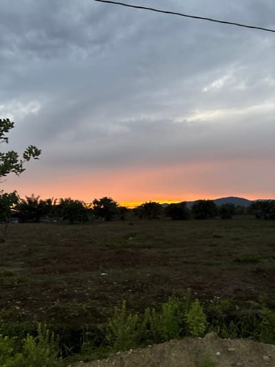 An image captured in Tela, Honduras, depicts a dusk or dawn sky over a rural landscape. The upper half of the frame is dominated by a grey, overcast sky, transitioning into a vibrant orange and yellow hue along the horizon line, indicative of a sunset or sunrise. Silhouetted against this bright horizon are distant, dark green mountains on the right, and a dense line of tropical vegetation, including various trees and what appear to be palm trees, extending across the midground. To the left, a partial structure is faintly visible behind the trees. The lower half of the image features an uncultivated field of dark earth with sparse green patches of grass and weeds. In the immediate foreground, at the bottom of the frame, there is an uneven mound of dirt with small green shrubs. A single dark utility wire traverses the upper third of the image from the upper left to the upper right. No individuals or active subjects are present in the scene.