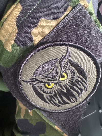 A close-up photograph shows a military-style patch attached to camouflage fabric, likely a uniform sleeve. The patch is round and features a stylized image of a bird of prey, possibly an owl or eagle, with sharp features and bright yellow eyes. The bird is rendered in black thread against an olive green background. The patch is secured to the camouflage fabric with a black Velcro-style backing. The camouflage pattern consists of a mix of green, brown, and black blotches. The location is not discernible from the image.