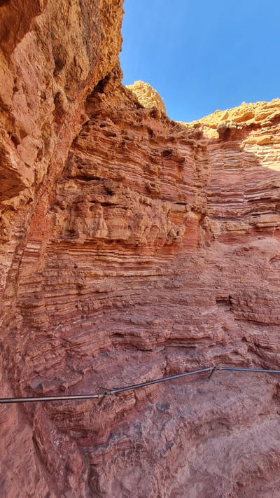 This photograph shows a close-up of a rust-colored rock formation with a mostly clear blue sky visible in the top left corner. The rock formation displays vertical striations, and there is a metal pipe with connectors attached to the rock face. The location is identified as being in the city of Hevel Eilot Regional Council, Israel. The scene does not contain any people, but the presence of the pipe suggests human interaction or alteration to the natural landscape.