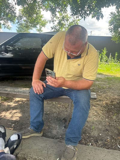 A middle-aged Caucasian man with a receding hairline and glasses is seated on a wooden bench outdoors, looking down at his smartphone. He is wearing a yellow polo shirt with a dark collar and sleeve trim, and blue jeans. The man is positioned in the center-right of the frame. To his left, a black car is partially visible, parked behind him. The background consists of a dark wooden fence, green vegetation, and trees with dappled sunlight filtering through their leaves. The ground is covered in dirt and dry grass, with some cigarette butts scattered around. In the foreground, the feet of two individuals are visible; one pair wearing black slides and white socks, the other wearing brown casual shoes. The scene suggests a casual outdoor setting, possibly a backyard or park area. The location is identified as Pereiaslavske, Ukraine.