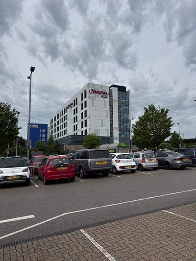 The image shows a parking lot scene in High Wycombe, United Kingdom. In the background is a modern multi-story building with the "Hampton by Hilton" logo displayed. Adjacent to it is a blue building marked "Access Self Storage." Several cars are parked in the foreground. Their number plates include: EY65 YBC, KR13 EUD, 3 RGR, RF17 AEE, DN10 WHC, and NX24 SKE. A street light pole is visible near the Access Self Storage building. The sky is overcast with dense clouds. The parking area is marked with white parking lines and features brick paving in certain sections.