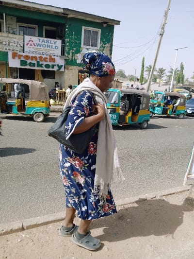A street scene in Mumbwa, Zambia, depicts a woman walking from right to left on a dirt path alongside an asphalt road. The woman wears a dark blue dress with a red and white floral pattern, a matching floral headscarf, a light beige fringed scarf draped over her left shoulder, and a black shoulder bag. She has grey clogs on over light-colored socks. Her shadow is cast to her right rear.
In the background, to the left, is a two-story building with peeling green paint. The ground floor displays signage for "Frentch Bakery" with orange and green lettering, and "TASLY WORLD" above it. A yellow and light-blue auto-rickshaw is partially visible near this building. On the asphalt road, several auto-rickshaws are present; one prominently displays "PTA LOCAL" on its side. A tall, lean utility pole with multiple wires extends upwards on the right side of the frame, under a bright, clear sky. Distant trees and another street light pole are visible further down the road. The environment suggests a busy, sunny urban setting.
