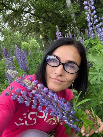 A woman with short black hair and blue eyes, wearing glasses and a pink sweatshirt, is in a field of lupines in Vakhrino, Russia. She is holding a bouquet of lupines. The background is filled with more lupines and green foliage. The woman is smiling and looking at the camera. The image is taken from a close-up perspective, focusing on the woman and the flowers.