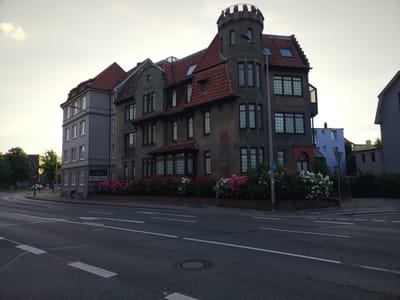 The image depicts a street view in Cuxhaven, Germany. In the foreground, a dark asphalt road with white lane markings is visible. To the right, a sidewalk and a grassy area with bushes and flowers line the road. In the background, a large, imposing building with a distinctive turret and red tile roof stands prominently. The building appears to be multi-story with numerous windows. Adjacent to it, another building with a gray facade and red tile roof is visible. Several other buildings, varying in color and architectural style, are present further in the background. The sky is overcast with a pale, diffused light, suggesting it may be early morning or late evening. A traffic light with a red signal is visible to the left, indicating potential traffic control at an intersection. No people are clearly visible in the scene.