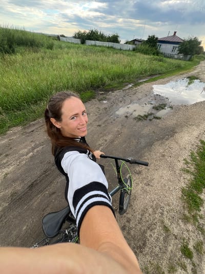 A woman rides a bicycle on a dirt road in Pivkino, Russia. The scene is rural, with a grassy field on the left and a house in the distance. The road is muddy with puddles, suggesting recent rain. The sky is cloudy, indicating overcast weather. The woman is smiling and looking at the camera, appearing happy and relaxed. She is wearing a black and white jacket with striped sleeves. The time of day appears to be late afternoon or early evening, given the lighting conditions.