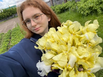 A young person with reddish-brown hair and fair skin, adorned with silver-framed glasses, is captured in a close-up, rotated outdoor selfie. They wear a navy blue long-sleeved top, possibly a sweatshirt, with a visible silver zipper pull. Their expression is neutral and calm as they gaze towards the camera.
The dominant feature of the image is a very large, vibrant bouquet of irises held by the person, filling a significant portion of the left frame. The flowers are primarily bright yellow, with some displaying delicate brown or light purple striations on their lower petals. A few white irises and hints of darker purple varieties are interspersed, adding variety to the large arrangement.
The setting appears to be an outdoor garden or rural area, consistent with a location like Новая Тура, Russia. The background features lush green grass and low-growing plants with small white flowers. Further back, dense green bushes and a rustic wooden fence are visible. In the distance, trees and what seems to be a building with a green roof are discernible under a bright, partly cloudy sky. The photograph was taken during daytime, likely morning or afternoon, indicated by the natural light. The image is oriented 90 degrees clockwise from a standard upright view. No text is visible.