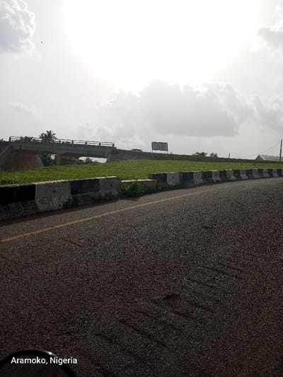 The image shows a paved road in the city of Iyin, Nigeria (latitude: 7.661, longitude: 5.167). The road curves to the left and passes under a concrete bridge. The road has a single yellow line down the middle. There is a grassy median separating the road from the grassy area on the right. The grassy area has a black and white curb. On the right side of the image, there is a white sign with red lettering that says "HOTEL." There are a few trees and bushes visible in the distance. The sky is cloudy with a bright white sun shining. There are no people or vehicles visible in the image.