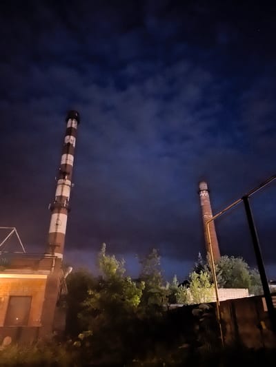 The image shows two industrial smokestacks against a dark, cloudy night sky. The smokestack on the left is taller and painted with alternating brown and white bands. It has visible numbers painted on it: "1989". Attached to its side are metallic structures. To the left of this smokestack is a brick building with a dark window and a section illuminated by a warm light. Trees and other vegetation are visible in the foreground and between the smokestacks. The smokestack on the right is shorter, made of brick, and has white markings. A metallic fence or railing runs along the right side of the image, partially obscuring the view of the right smokestack and some brick structures behind it. The scene appears to be within or adjacent to an industrial area in Ново-Талицы, Russia.
