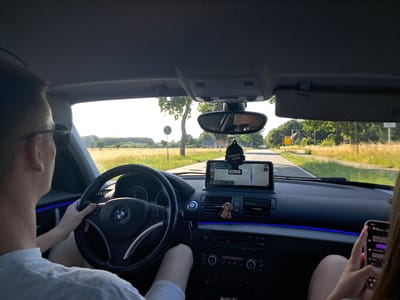 This media file captures an interior view of a car driving on a road in Friedrichsruhe, Germany. The primary subjects are two people: the driver, a man wearing sunglasses, and a passenger, whose hands are visible holding a smartphone. The scene is set within the car's cabin, looking forward through the windshield.

The activity is driving, with the driver's hands on the steering wheel of a BMW. A navigation system is displayed on a screen mounted on the dashboard, showing a route with the destination "Black Ice" and a distance of 20 km. A black air freshener with the text "WUNDER-BAUM" hangs from the rearview mirror. A small, plush monkey toy is also visible on the dashboard, near the air vents.

Notable details include the time of day, which appears to be afternoon or early evening, suggested by the warm, golden light illuminating the landscape. The weather seems clear and sunny, with bright daylight. The setting is a rural or semi-rural area, with fields of dry grass and trees lining the road, and a few houses visible in the distance. The interior of the car features ambient blue lighting along the dashboard. The passenger is actively engaged with their smartphone, suggesting a casual or leisurely drive. No explicit emotions are conveyed, but the overall impression is one of a relaxed journey.