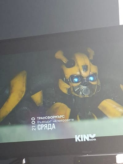 The image shows a television screen displaying a promotional advertisement for the film "Transformers: Rise of the Beasts" on Kino Nova channel. The ad features a close-up of the Transformer Bumblebee's head. Text on the screen, written in Bulgarian, indicates that the movie will air on Wednesday at 21:00 (9:00 PM). The photo was taken indoors. There is no indication of Selyanin or Bulgaria specifically.