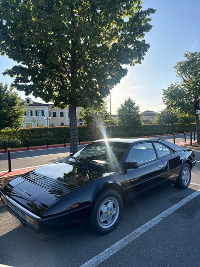 In Serravalle Scrivia, Italy, a black Ferrari Mondial T is parked in a lot adjacent to a road. The car is positioned in a parking space marked with a white line. Behind the car, a manicured hedge runs along the edge of the road. Beyond the hedge, a building with a light yellow facade, featuring black shutters and a decorative architectural element on the roof, is visible. Several trees with green foliage are scattered throughout the scene, with one prominently positioned to the left of the car and another to the right. The sky is clear blue, with the sun casting a bright glare on the car and through the trees. The overall environment appears to be an outdoor area, possibly near a commercial or residential complex.