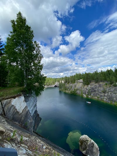 This image depicts a scenic view of the Ruskeala Mountain Park, a former marble quarry in Ruskeala, Russia. The foreground is dominated by a steep, rocky cliff face with visible layers of white and grey marble. A large birch tree with white bark and green foliage grows precariously from the edge of the cliff, its branches extending over the water.
The middle ground features a serene, deep blue lake, likely a flooded part of the quarry. The water is clear, with visible underwater features and a few yellow buoys forming a line across the surface. A small white boat with red trim is visible on the lake. The surrounding landscape is characterized by rugged, grey rock formations that form the steep banks of the quarry.
In the background, a dense forest of green coniferous and deciduous trees covers the hills. A narrow walkway or path is visible along the top of the quarry wall, with some structures or viewing platforms visible. Further in the distance, a tunnel entrance can be seen carved into the rock face, hinting at the historical use of the quarry for marble extraction. The sky above is a vibrant blue, dotted with fluffy white clouds, suggesting a clear and pleasant day. The overall impression is one of natural beauty and historical significance, showcasing the transformation of an industrial site into a popular tourist destination.