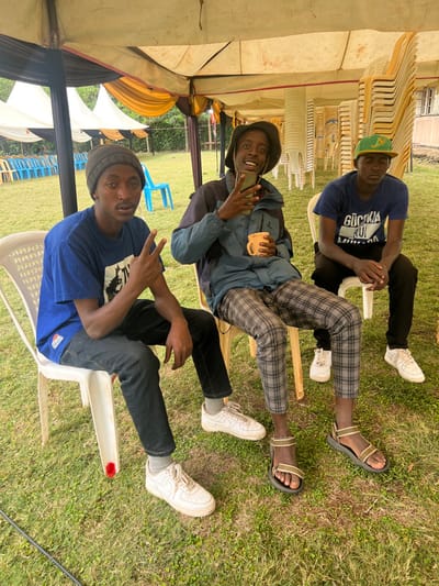 The image, taken in Makuyu, Kenya, shows three young men sitting on white plastic chairs outdoors under a large tent during daytime. The ground is covered with grass, and stacks of similar chairs are visible in the background, suggesting an event setting.
From top to bottom in the upright image orientation:
The man at the top wears a blue t-shirt with the text "GÜCKNA RUI MUMBA" printed across the front, black pants, and a green and yellow baseball cap. He appears to be resting with his eyes closed, wearing white sneakers.
The man in the middle wears a blue jacket over a t-shirt, distinctive plaid trousers, and a dark bucket hat. He holds a smartphone in his left hand and a yellow mug in his right, smiling brightly at the camera. Partial text "GUCCIA RUI..." is visible on his t-shirt. He wears beige and black sandals.
The man at the bottom wears a blue t-shirt, dark jeans, and a dark beanie. He is also smiling at the camera and making a peace sign with his right hand. He wears white sneakers.
The scene conveys a relaxed and casual atmosphere under natural daylight.