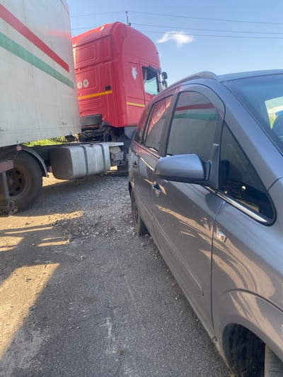 A gray car is parked on the right side of the image, facing away from the viewer. To the left of the car is a red semi-truck, also facing away from the viewer. Between the car and the truck is a large white trailer, which appears to be connected to the red semi-truck. The trailer has three horizontal stripes: green, red, and green from top to bottom. The ground is a mixture of asphalt and gravel. The sky is blue with a few clouds. The scene appears to be in an outdoor, possibly industrial or parking lot, area. The image was taken in Mais'ke, Ukraine.