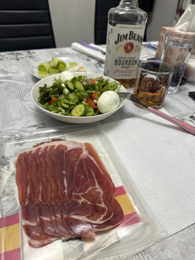 This image displays a domestic dining setup on a table in the city of Lancaster, United Kingdom. No individuals are present. In the foreground, a vacuum-sealed plastic package contains numerous slices of cured ham, characterized by a reddish-brown color and visible fat marbling. The package features red and yellow striping at its lower edge. Centrally positioned, a white bowl holds a fresh salad comprising green leafy greens, sliced cucumbers, halved cherry tomatoes, and two whole, peeled hard-boiled eggs. A silver fork with a pink handle is partially submerged in the salad. To the right of the salad, a partially full clear glass bottle of "Jim Beam Kentucky Straight Bourbon Whiskey" stands upright. Adjacent to the bottle, a clear glass tumbler with a decorative emblem contains an amber liquid, consistent with whiskey. In the background, a small white plate displays slices of blanched leeks and another indeterminate food item. Further to the right, a white tissue box featuring a rainbow motif and a clear glass with a similar rainbow pattern are visible. The table surface is covered by a light-colored, patterned tablecloth. A dark, possibly black, upholstered chair back is partially visible in the distant background.